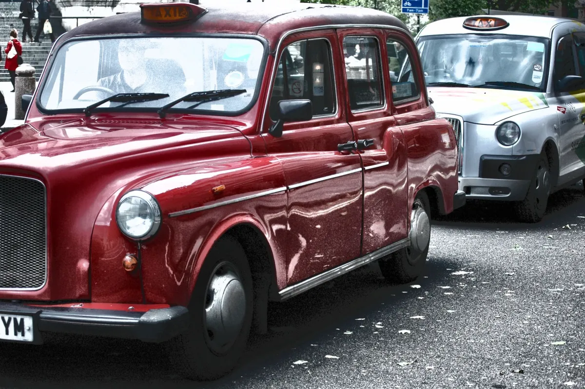 Londra'da bir caddede park etmiş kırmızı klasik bir taksi ve arkasında gri bir taksi